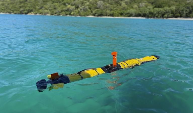 The yellow, cylindrical IVER3-AUV floats in clear blue-green water.